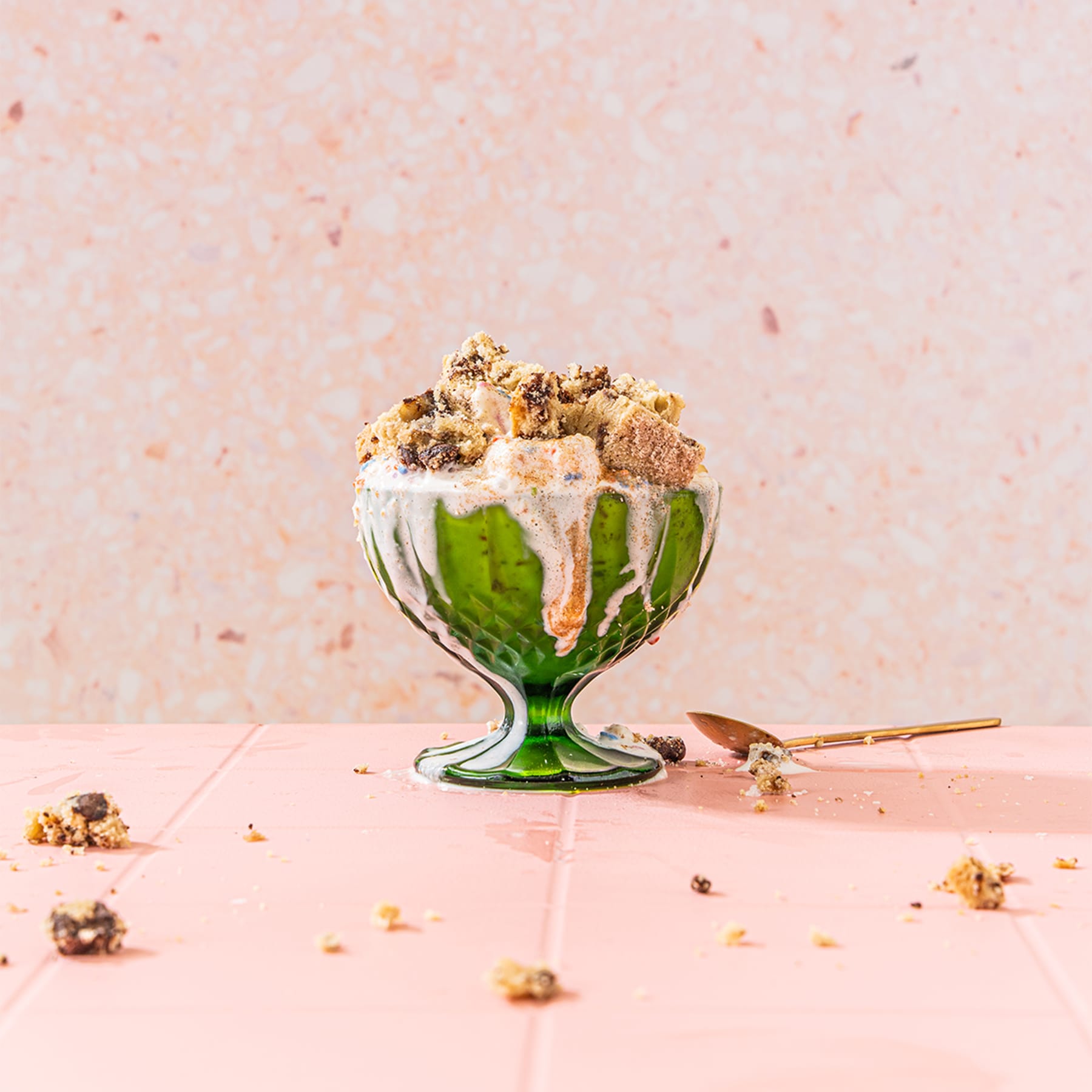 Wandel bites sit in a green glass pedestal bowl on top of melted ice cream with a spoon and additional bite crumbs on the table below with pink background.