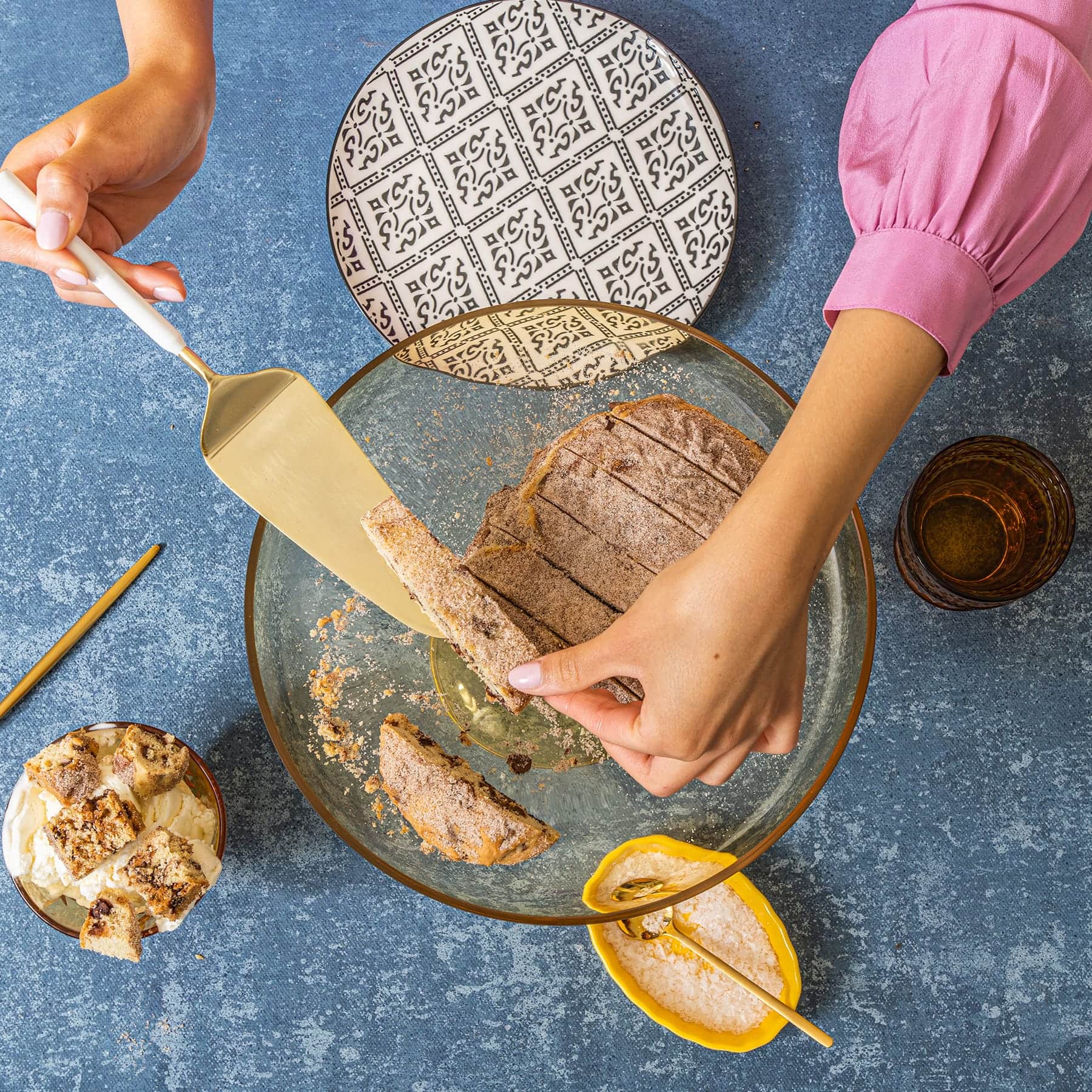 A woman's hands in a pink shirt lift a slice of Wandel loaf from a clear glass bowl with a black and white print plate, a glass of water, a small tray of salt with spoon, and a small bowl of wandel bites sit around the plate.