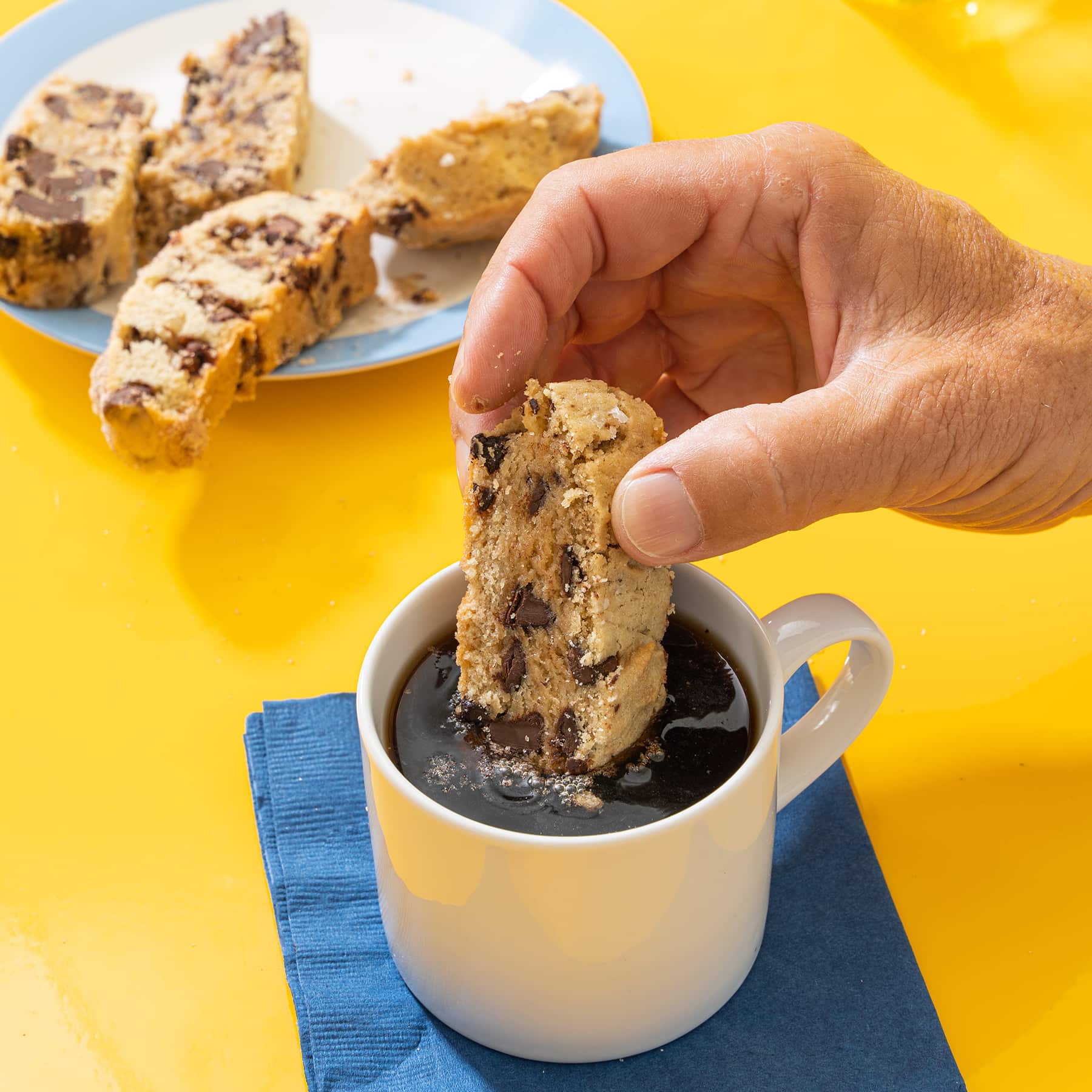 A gentleman's hand dips wandel bread slice into a cup of coffee with additional slices on plate in background.