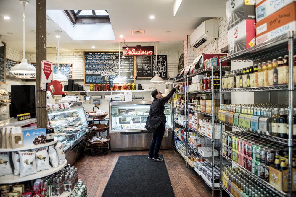 Shelky's inner store photo with metro racks with product on the right, a delicatessen counter and side at the center, and a ticket pull down on pole to left.