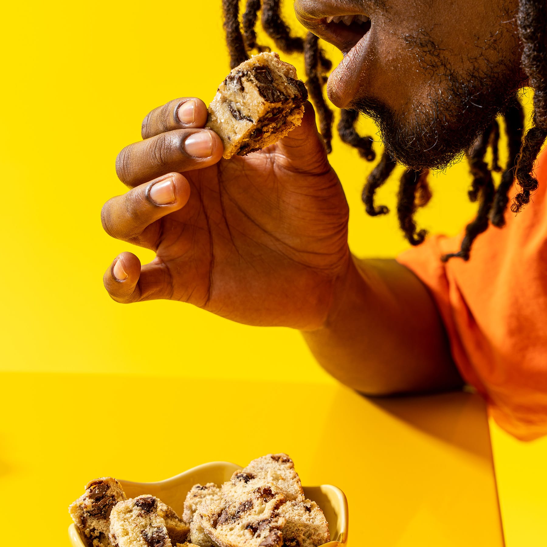Bright yellow background offsets gentlemen bringing a dark chocolate wandel bite to his mouth from a bowl of bites below.