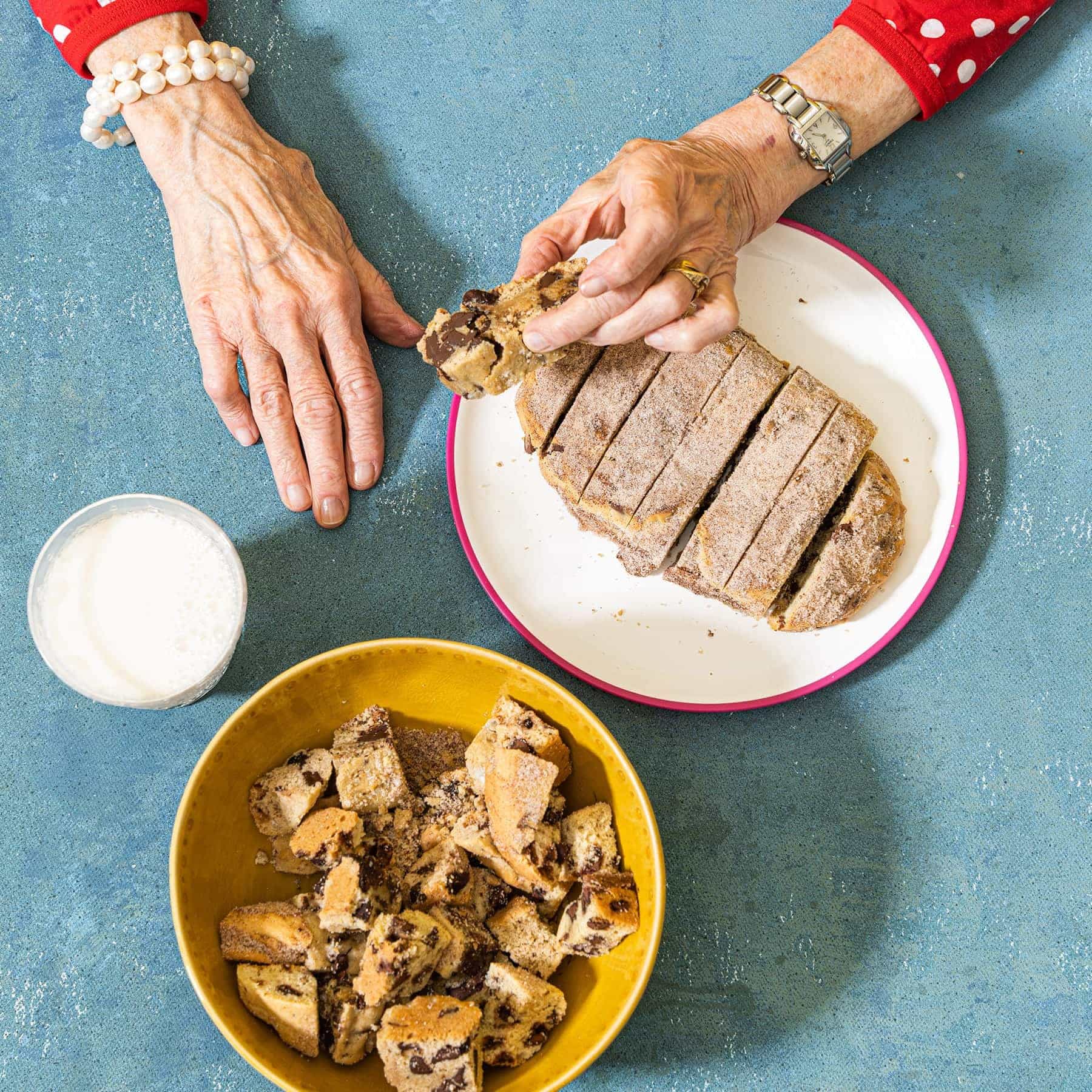 Grandmother's hands in red and white polka dot shirt and pearl bracelet holding a slice of Wandel loaf on plate. Additional bowl of dark chocolate wandel bites and milk nearby.