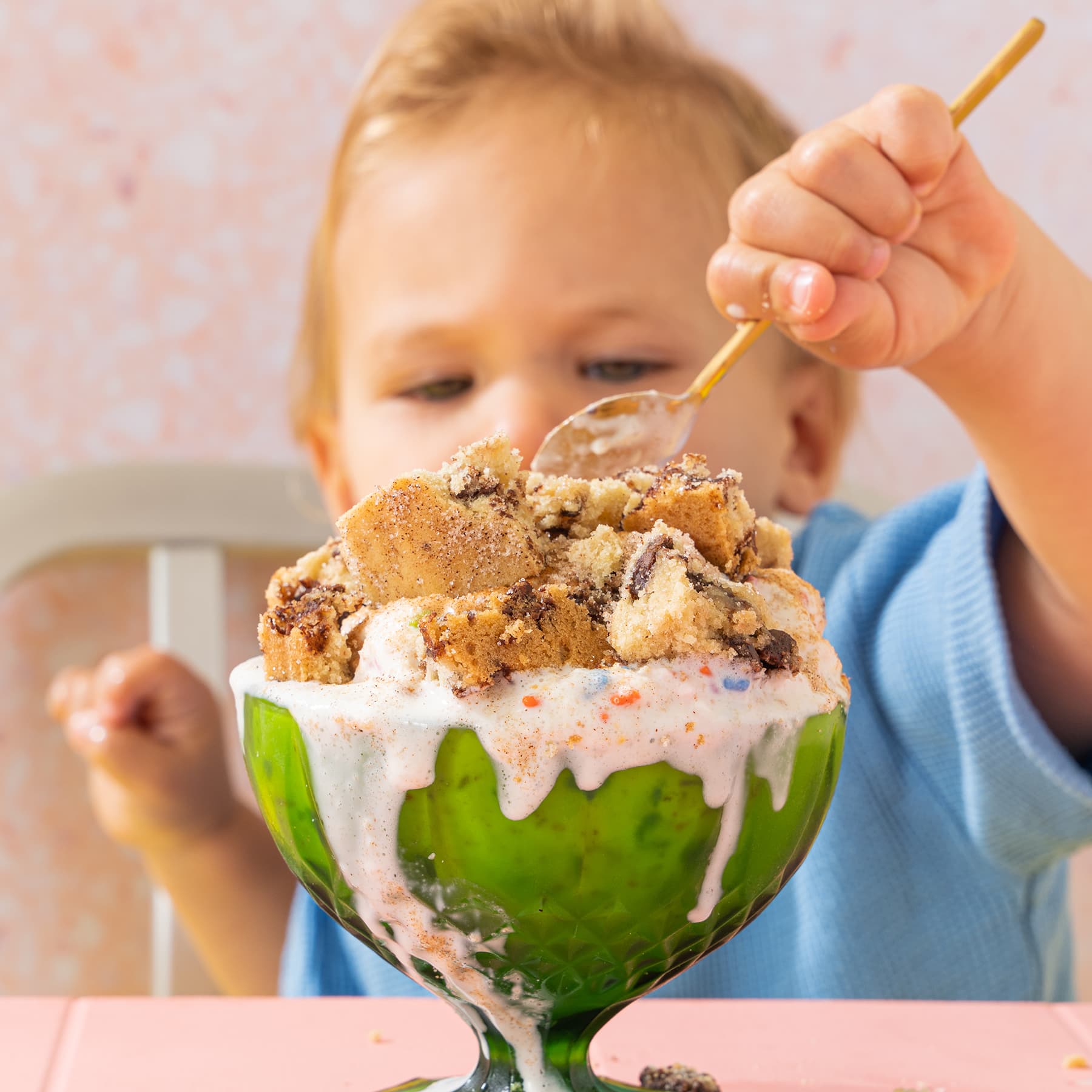 Small boy uses a spoon to dig into Wandel bites in a green glass pedestal bowl on top of melted ice cream on pink table.