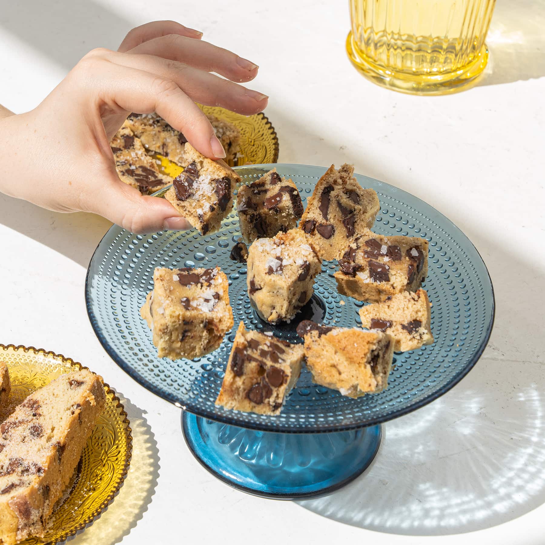 Dark chocolate + sea salt wandel bites on blue glass pedestal plate with hand selecting a piece, and assorted yellow glassware in background